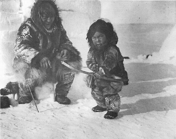 Un hombre inuk enseña a un niño a disparar un arco y una flecha, alrededor de 1920.