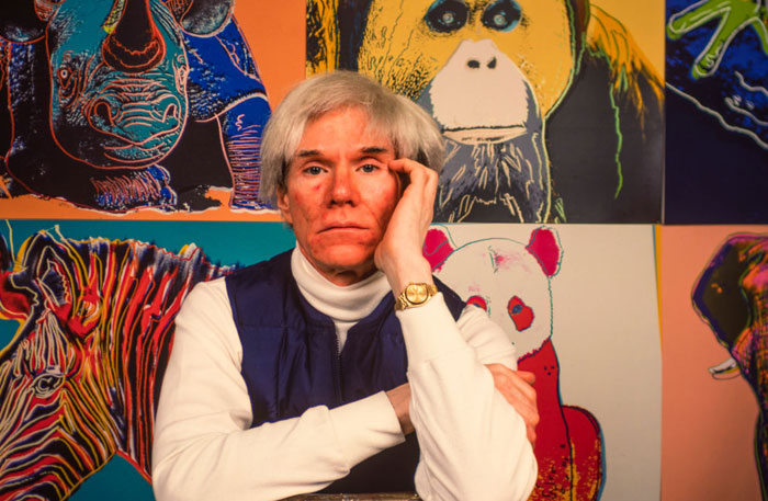 A portrait of the famous artist Andy Warhol, resting his head on his hand, with vibrant animal artwork in pop art style behind him.