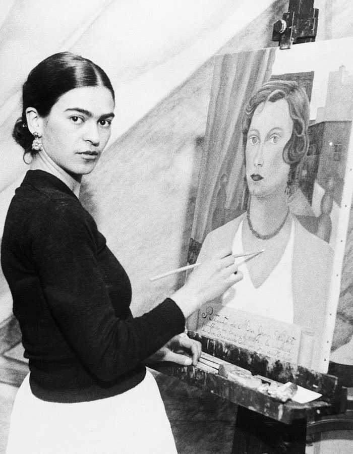 A black-and-white photograph of the famous artist Frida Kahlo, painting a portrait at an easel, looking toward the camera.