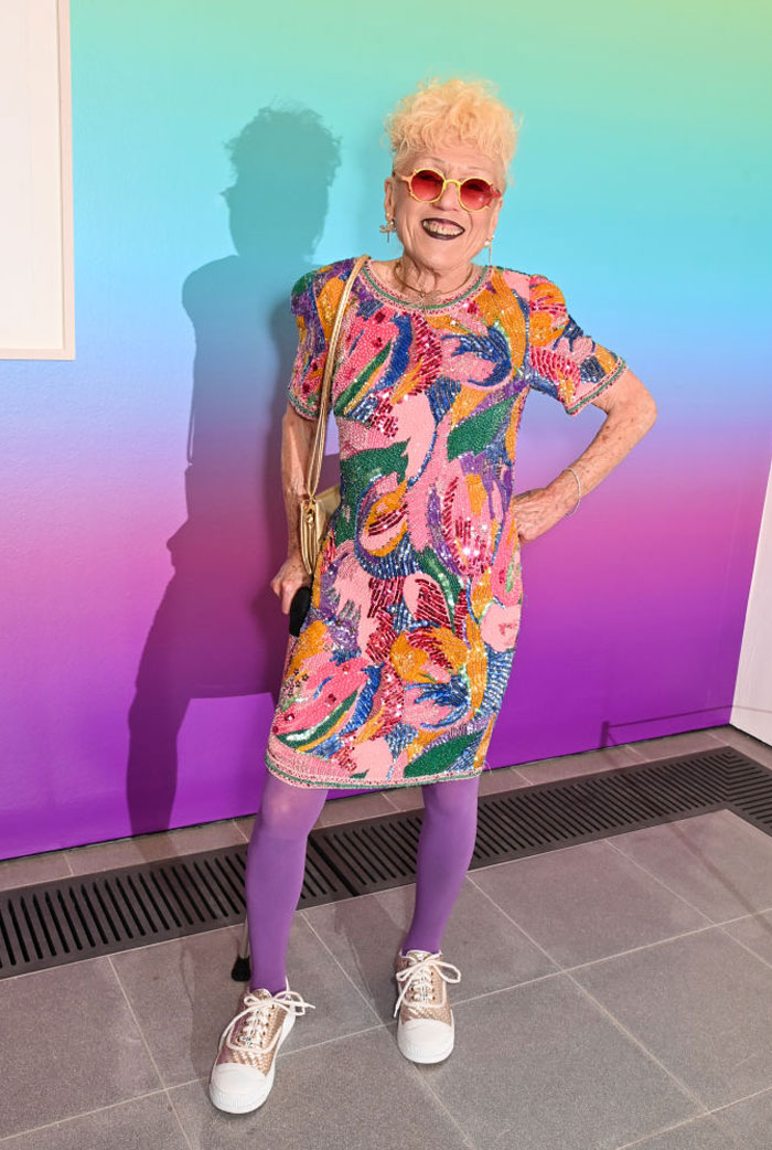 Judy Chicago, the famous artist, wearing a colorful, sequined dress, purple tights, and sunglasses, standing against a gradient background from blue to pink.