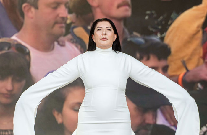 Marina Abramović, the famous performance artist, wearing a white dress with extended sleeves, standing in front of a crowd during a performance.