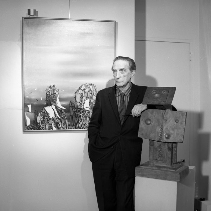 A black-and-white photo of the famous artist Marcel Duchamp, standing beside a sculpture and a painting, wearing a suit and looking contemplative.