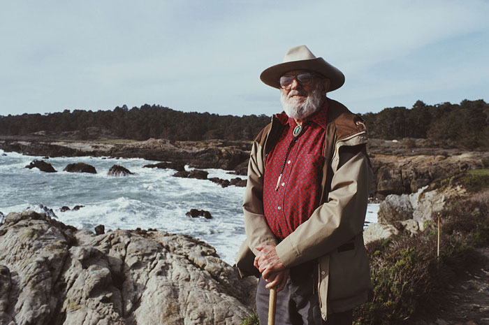 The famous artist Ansel Adams, wearing a hat and a red shirt, standing with a cane by a rocky shoreline with waves crashing behind him.