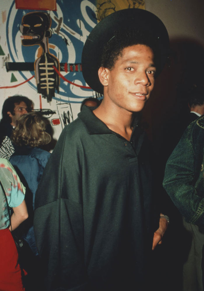 A candid photo of the famous artist Jean-Michel Basquiat, wearing a black hat and dark clothing, standing in front of one of his graffiti-inspired artworks.