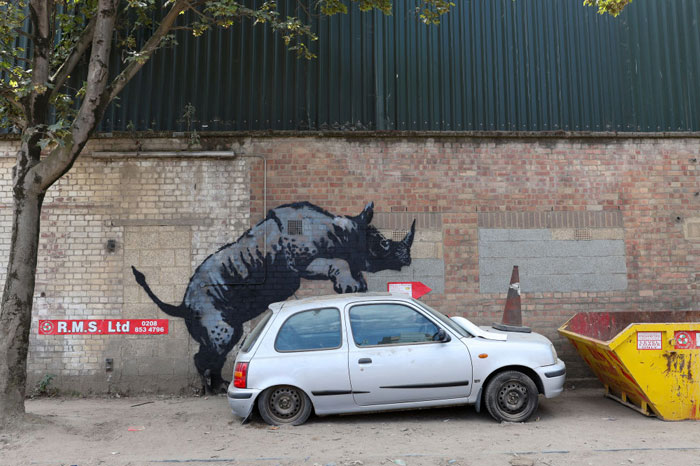 A street artwork by the famous artist Banksy, depicting a rhino stenciled on a brick wall behind a damaged car, creating an illusion of the rhino pushing the car.