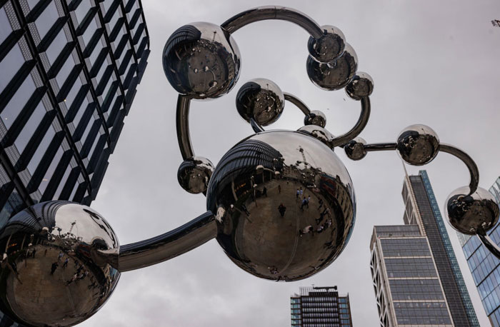 A large reflective sculpture resembling a molecular structure, by a famous artist Yayoi Kusama , displayed against modern buildings and a cloudy sky.