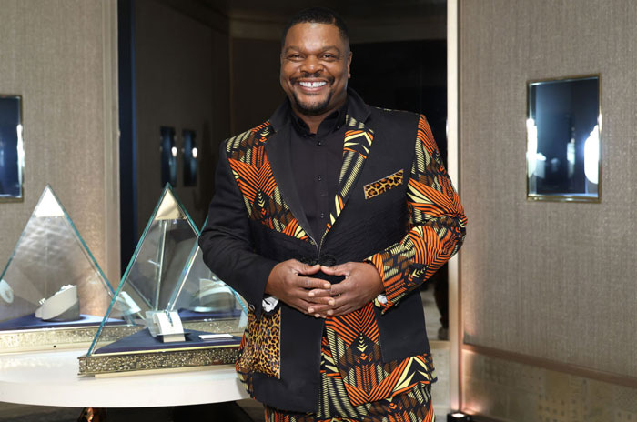 The famous artist Kehinde Wiley, smiling and wearing a vibrant, patterned suit, stands in an art gallery or exhibition space with glass display cases behind him.