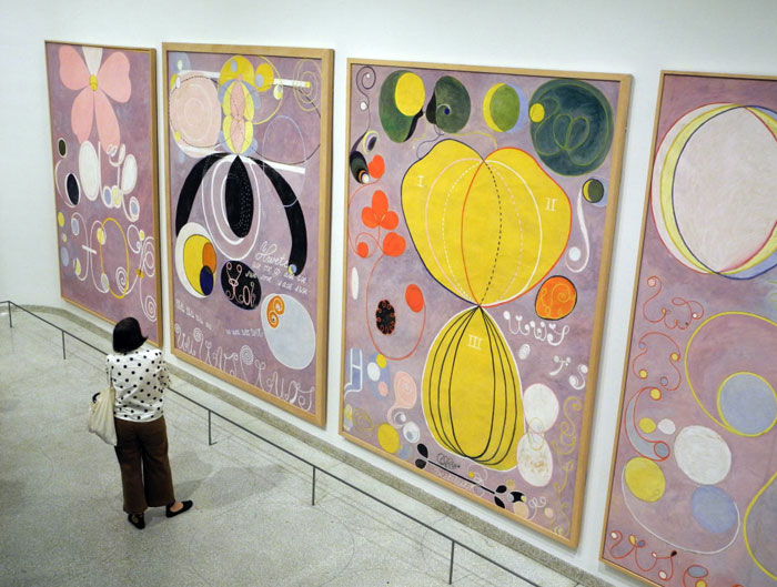 A visitor observes large, abstract paintings by the famous artist Hilma af Klint, featuring colorful shapes, spirals, and symbols on pastel backgrounds.