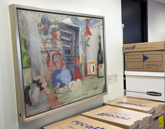 A framed abstract painting by a famous artist Alma Thomas hangs on a wall, next to stacks of cardboard storage boxes labeled "recall" in an office setting.