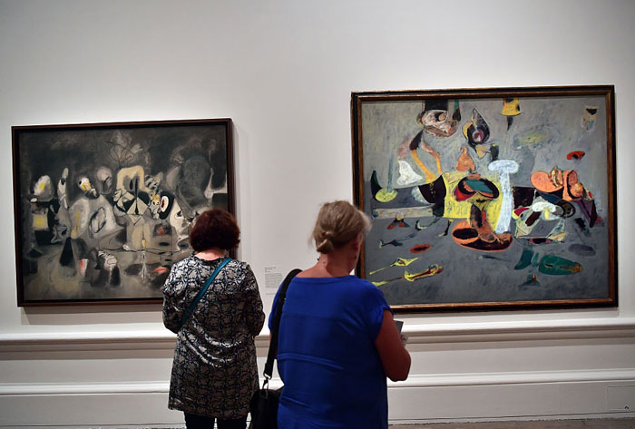 Visitors observe abstract paintings by the famous artist Arshile Gorky in a gallery, featuring surreal shapes, muted colors, and imaginative compositions.