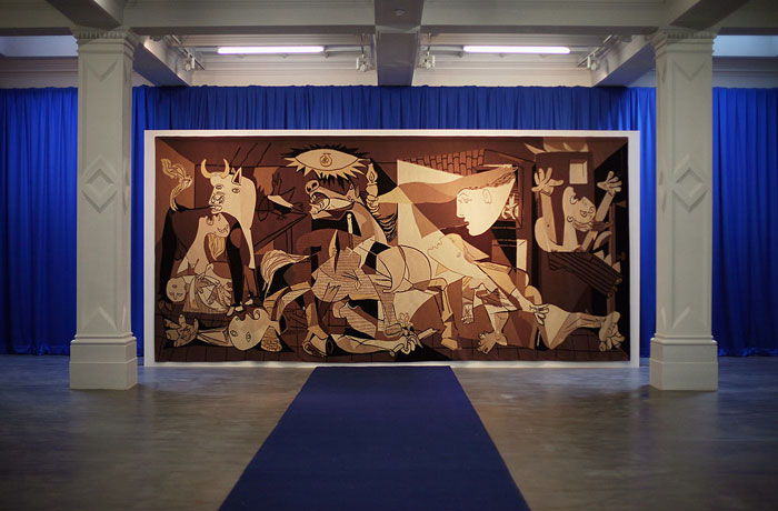 A large-scale reproduction of the famous artist Pablo Picasso's painting Guernica, displayed in a gallery with blue curtains and a blue carpet.