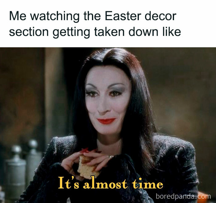 A Halloween meme featuring a character with long black hair and red lipstick, smiling mischievously while holding food. The text above reads, "Me watching the Easter decor section getting taken down like," and below it says, "It's almost time," referencing the anticipation for Halloween.