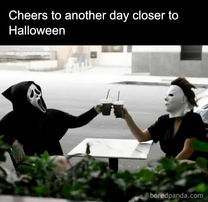 A Halloween meme showing two people in horror movie masks, one dressed as Ghostface and the other as Michael Myers, sitting outside at a table and toasting coffee cups. The text above reads, "Cheers to another day closer to Halloween."