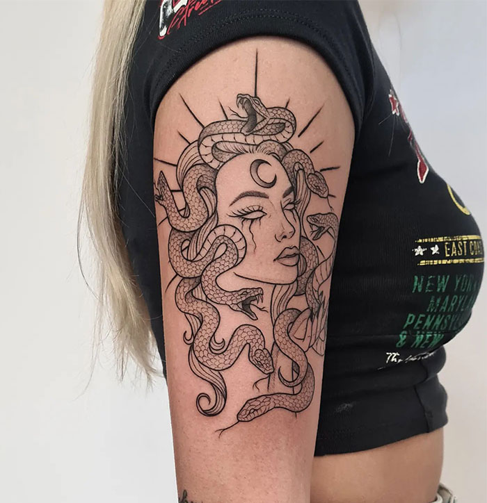 A minimalist black line Medusa tattoo on the upper arm, featuring a woman's face with snakes in her hair, a crescent moon on her forehead, and rays emanating behind her head.