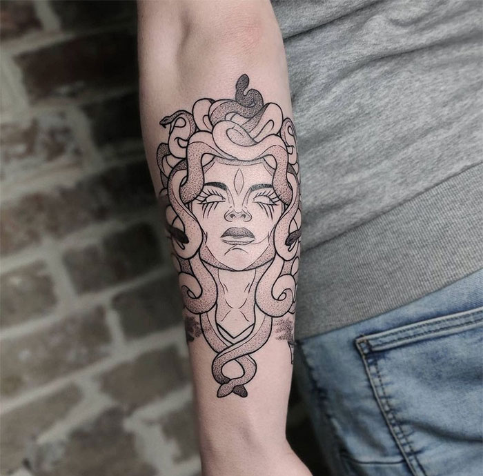 A Medusa tattoo on the forearm featuring a woman's face with closed eyes and snakes in her hair, drawn in bold black lines with subtle shading and intricate details.