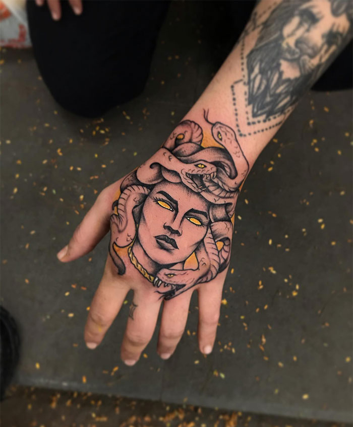  A Medusa tattoo on the hand featuring a woman's face with glowing yellow eyes, snakes coiling around her head, and detailed shading in dark and golden tones.