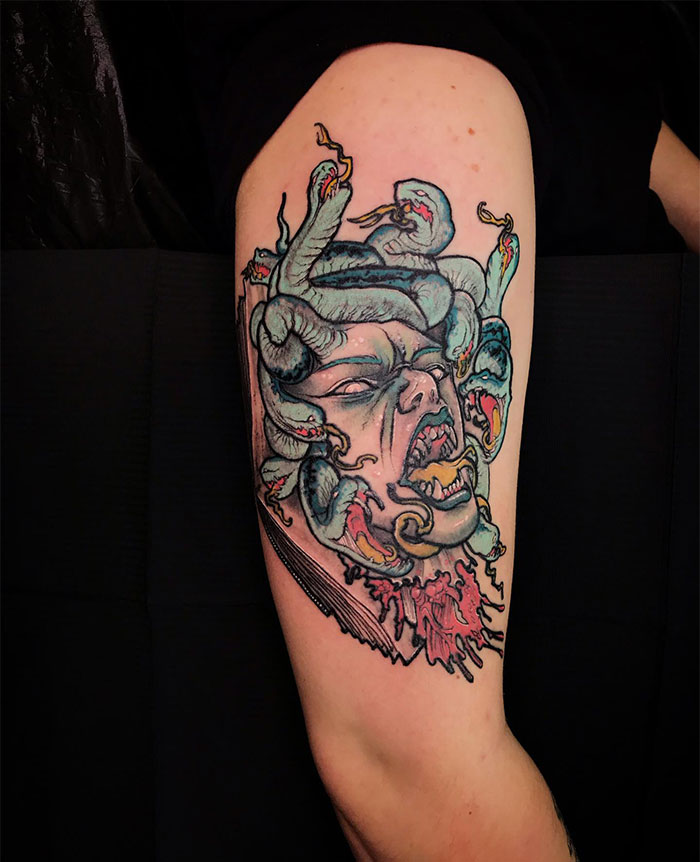 A colorful Medusa tattoo on the upper arm, featuring a fierce, screaming face with snakes in her hair, intense shading, and vibrant details, including blood dripping from the neck.