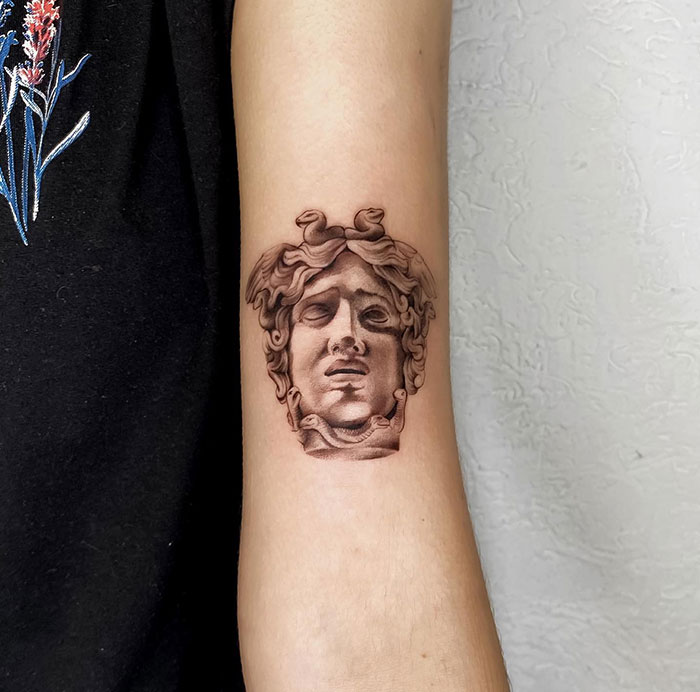 A realistic Medusa tattoo on the forearm, depicting a detailed stone-like bust of Medusa's face with intricate shading, capturing a classical sculpture style.