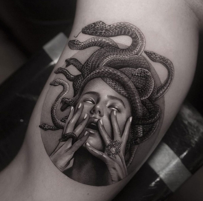  A realistic black and grey Medusa tattoo featuring a woman's face in an expressive pose, with detailed snakes coiling around her head, and her hands dramatically placed on her face.