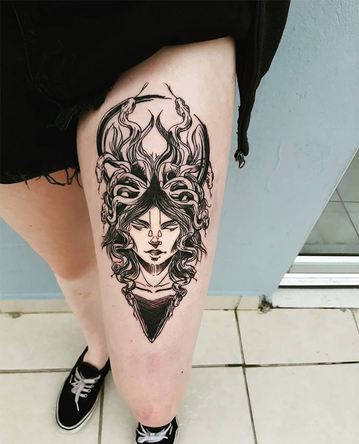 A black ink Medusa tattoo on the thigh, featuring a woman's face with intense eyes and snakes rising dramatically from her head, drawn in a detailed and bold line art style.