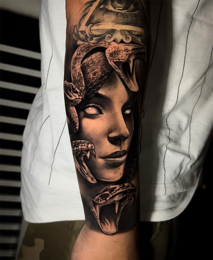 A realistic black and grey Medusa tattoo featuring a woman's face with glowing white eyes, surrounded by detailed snakes, some with open mouths, and intricate shading throughout.