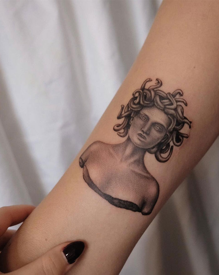 A realistic Medusa tattoo on the forearm, featuring a bust-like design with intricate shading, showing a classical sculpture style with snakes in her hair and detailed textures.