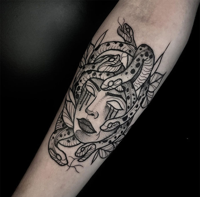 A black and grey Medusa tattoo featuring a woman's face with snakes intertwined in her hair, her eyes closed, and tears streaming down her face, surrounded by detailed leaves and intricate shading.