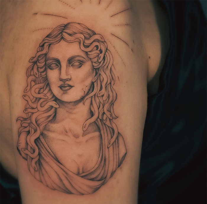  A detailed Medusa tattoo depicting a classical bust with snakes subtly coiled in her hair, sculptural shading, and fine lines radiating from her head, resembling ancient artwork.