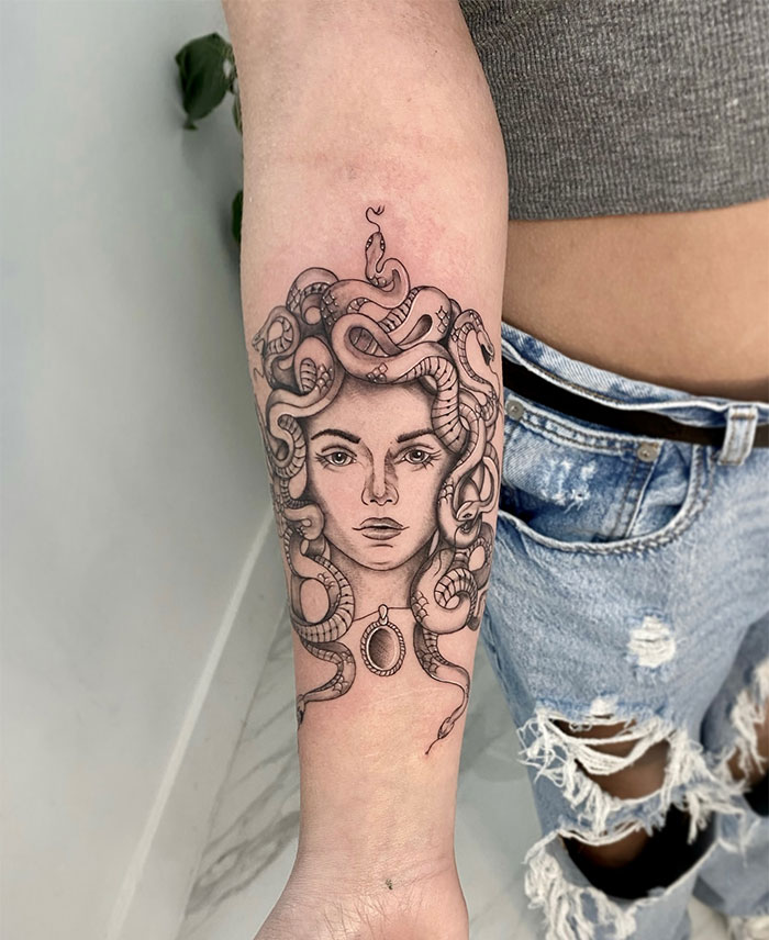 A detailed black and grey Medusa tattoo on the forearm, featuring a woman's face with snakes entwined in her hair, and a pendant hanging below her neck.