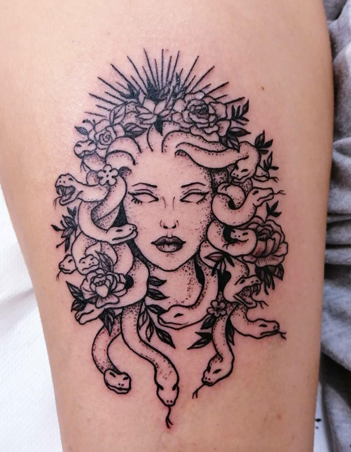 A black and grey Medusa tattoo featuring a woman's face with blank eyes, surrounded by snakes and flowers, with rays emanating from the top of her head.