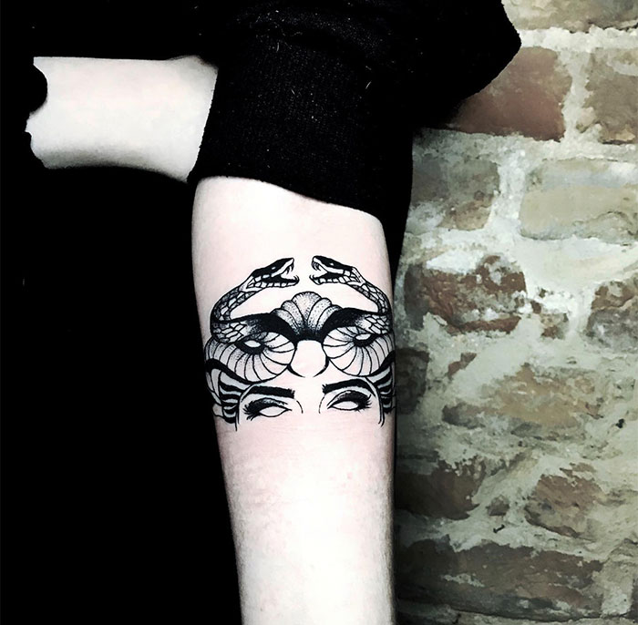 A minimalistic Medusa tattoo on the forearm, featuring only the upper part of a woman's face with two detailed snakes intertwined above her eyes.