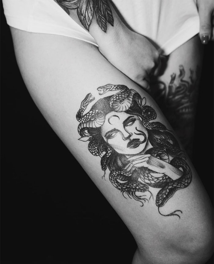  A black and grey Medusa tattoo on the thigh, featuring a woman's face with closed eyes, snakes coiling in her hair, and a crescent moon on her forehead.