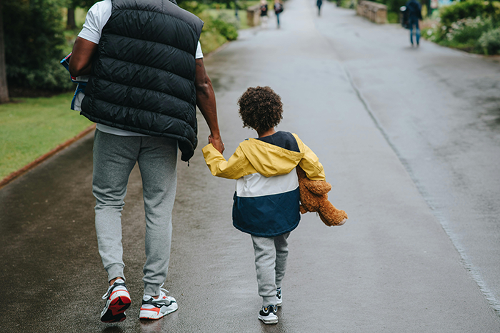 Child holding teddy bear, walking with an adult on a rainy path.