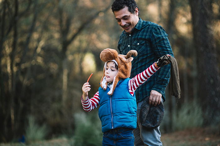 Child in a playful hat with an excited gesture, accompanied by an older man in a forest setting.