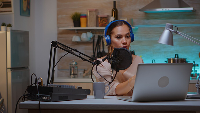 Woman recording a podcast at home, wearing blue headphones, speaking into a microphone, with a laptop open in front.