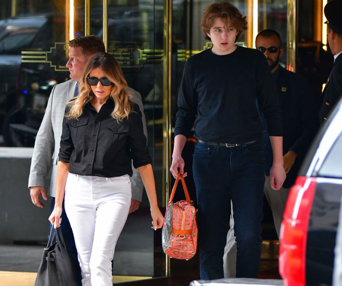 Barron Trump, noticeably taller, walks beside Melania Trump, wearing casual black and jeans, further highlighting Barron Trump's height.