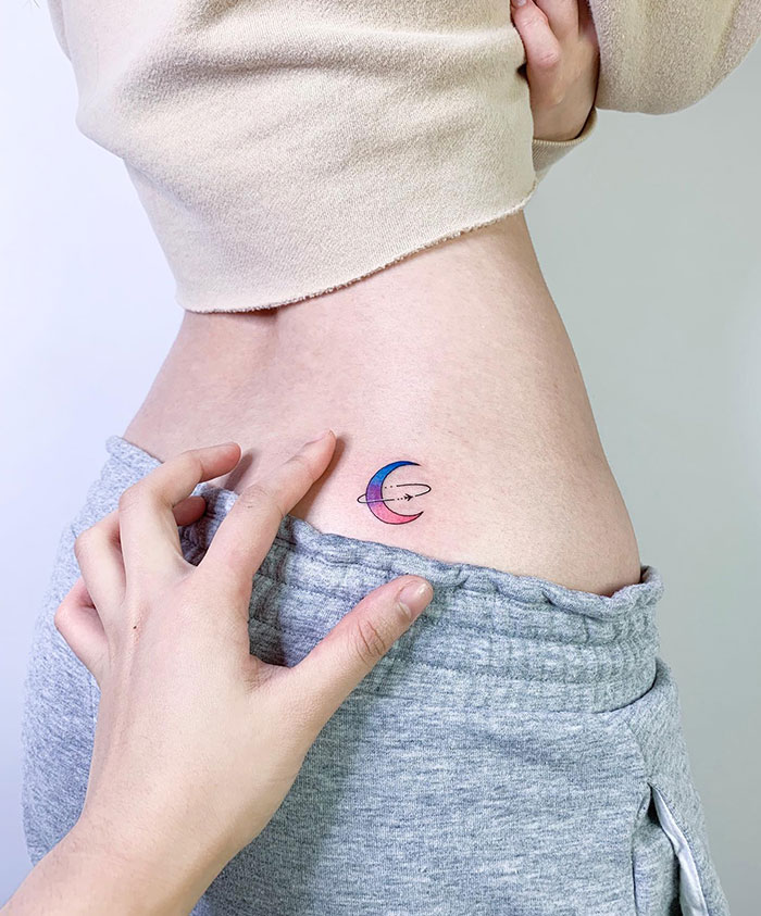 "A small tattoo idea of a colorful crescent moon with a minimalist planet ring design on the side of the torso, adding a cosmic touch."