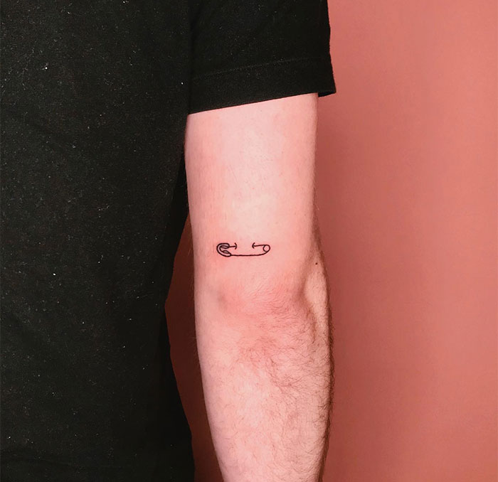 "A small tattoo idea of a simple outline of a safety pin on the back of the upper arm, adding a minimalist and edgy touch."