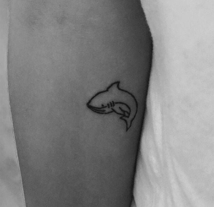 "A small tattoo idea featuring a simple black outline of a smiling shark on the upper arm, adding a cute and bold touch."