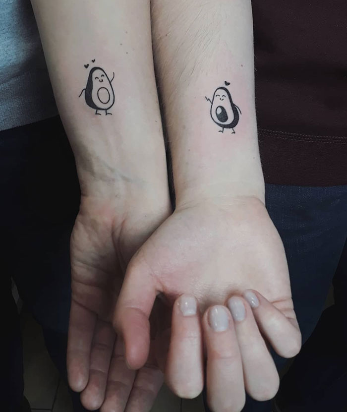 "A small tattoo idea featuring matching cute avocado characters on each wrist, symbolizing friendship or love, with tiny hearts above."