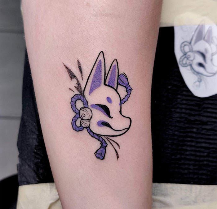  "A small tattoo idea featuring a stylized Japanese fox mask with purple accents and a decorative rope, symbolizing cultural and mystical themes."