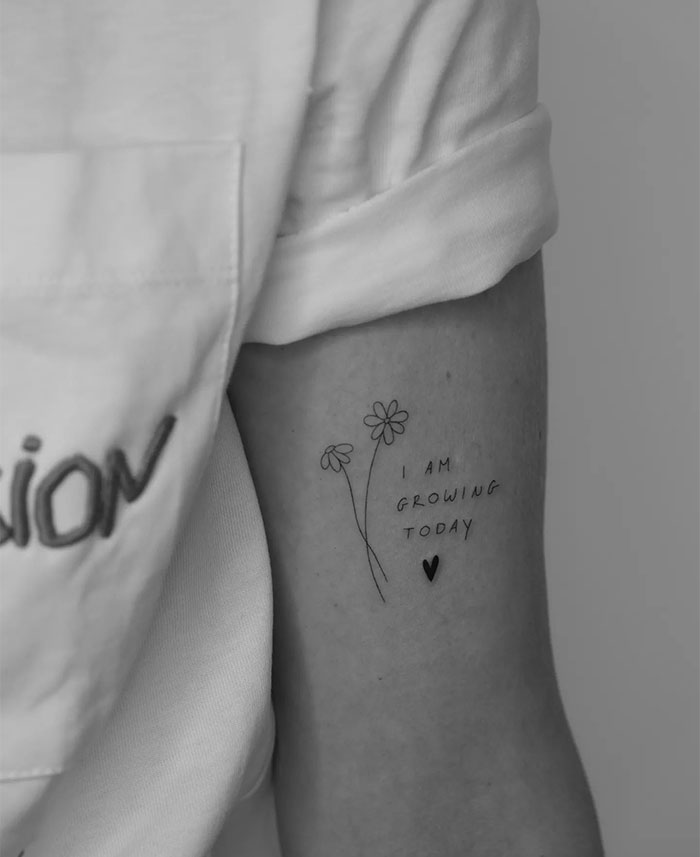 A small tattoo idea featuring two minimalist flowers and the words "I am growing today" with a small heart symbol, placed on the upper arm.