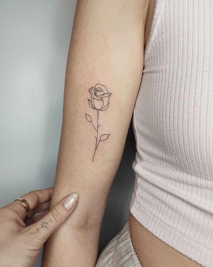 A small tattoo idea featuring a simple, minimalist outline of a rose on the inner forearm, with delicate leaves on the stem.