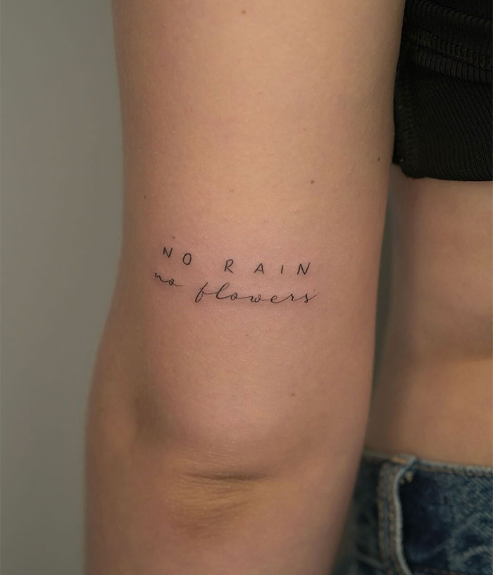 A small tattoo idea featuring the phrase "No rain, no flowers" in delicate cursive and uppercase lettering on the back of the arm.