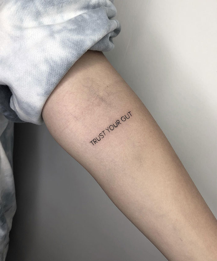 A small tattoo idea featuring minimalist text reading "TRUST YOUR GUT" in black ink on the inner forearm.