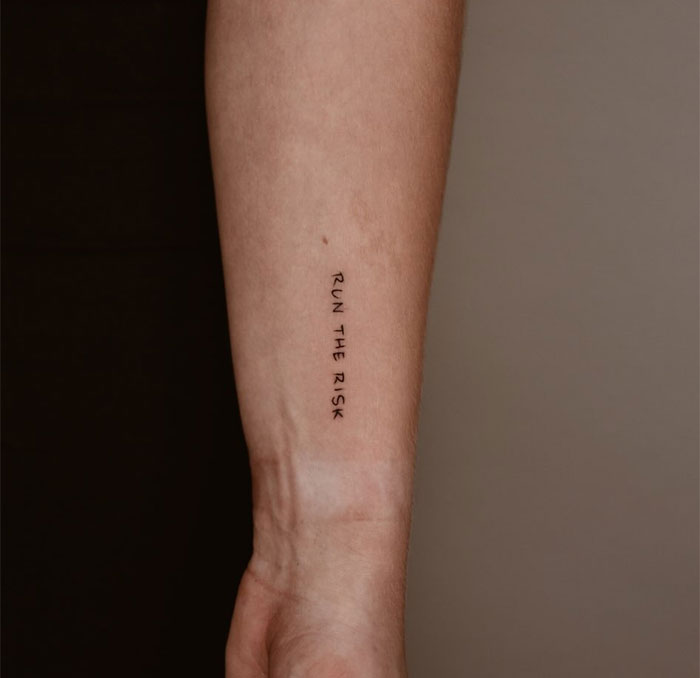 A small tattoo idea featuring the phrase "RUN THE RISK" in simple, vertical black text along the forearm, inspiring boldness and the courage to take chances.