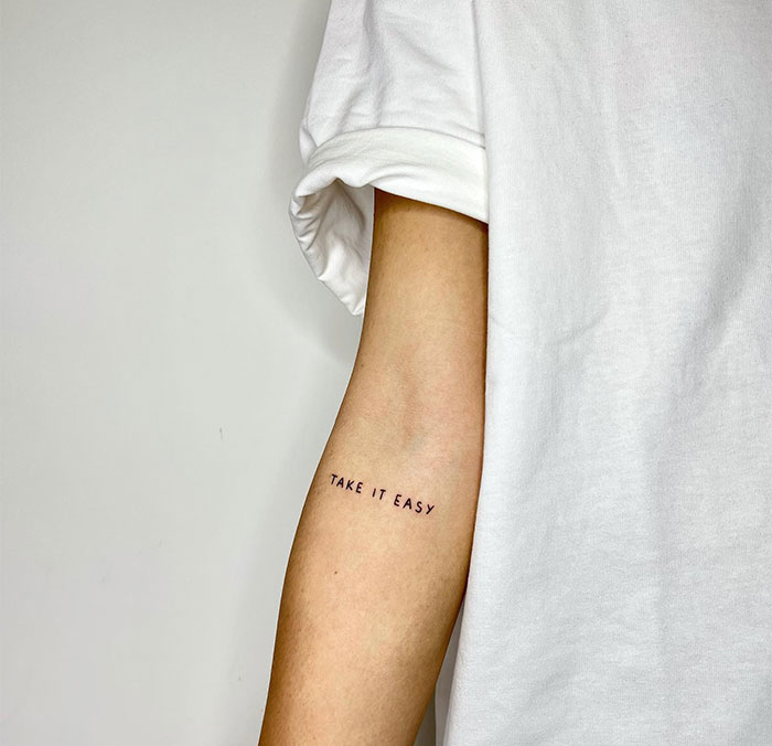 A small tattoo idea featuring the phrase "TAKE IT EASY" in a simple uppercase font on the inner arm, serving as a gentle reminder to stay calm and relaxed.