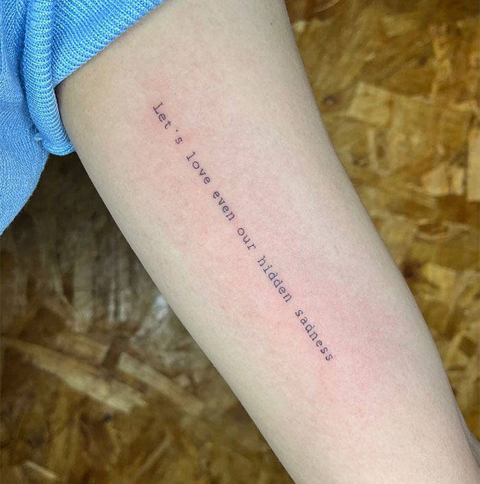 A small tattoo idea featuring the phrase, "Let's love even our hidden sadness," in a simple, typewriter-style font along the inner forearm, reflecting themes of self-acceptance and compassion.