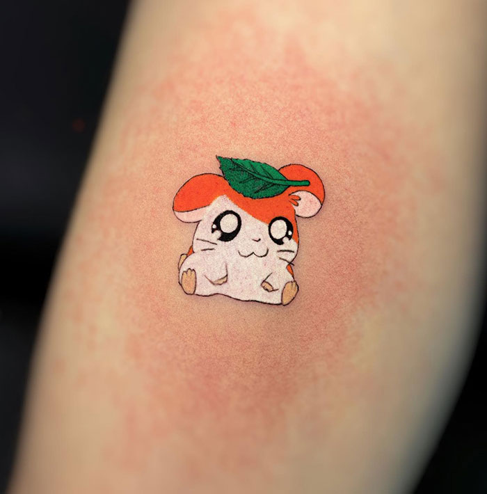 A small tattoo of a cute character with big eyes, orange ears, and a green leaf on its head, adding a playful and charming touch.