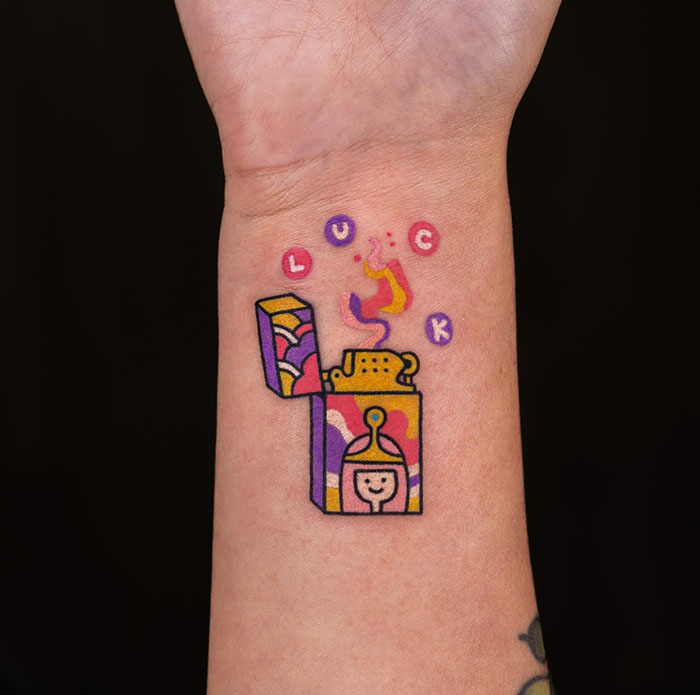 A small tattoo featuring a colorful, stylized lighter with the word "LUCK" floating above, creating a playful and vibrant design.